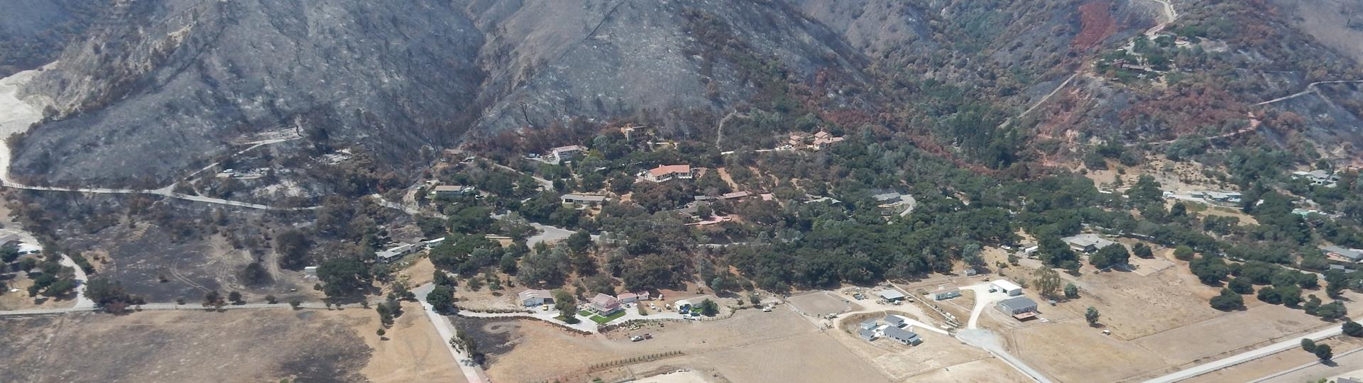 Mountains denuded by wildfire loom over homes on flat land below. The figure caption provides more information.