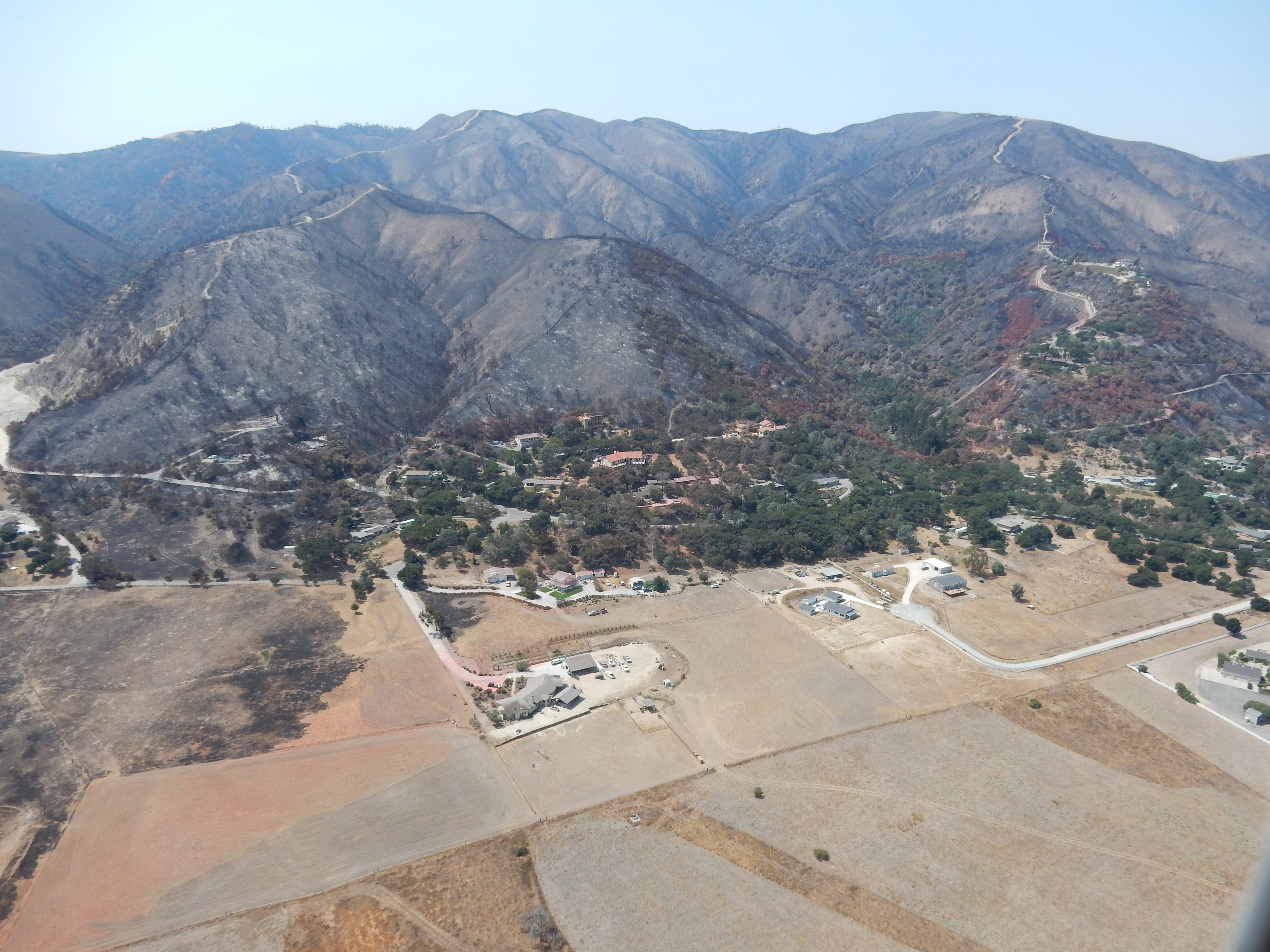 Mountains denuded by wildfire loom over homes on flat land below.