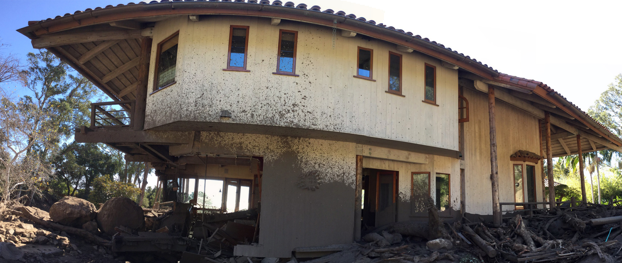 A two-story home damaged by a debris flow. The building is surrounded by displaced boulders and woody debris, and the exterior is caked with mud up to the bottom of the second floor. The caption below provides more information.