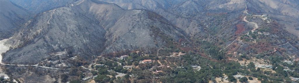 Mountains denuded by wildfire loom over homes on flat land below. The figure caption provides more information.