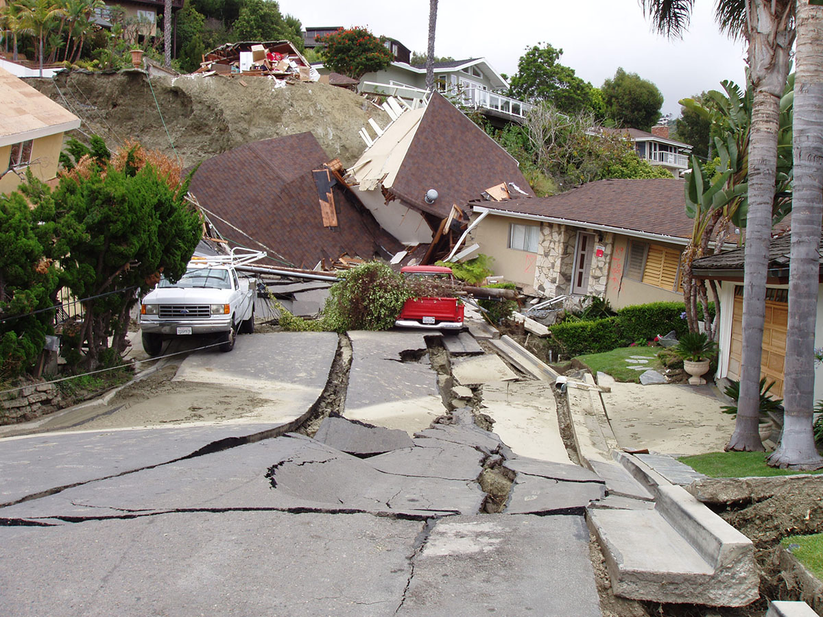 Photograph of homes along Flamingo Drive dropped down by the June 2005 Bluebird Canyon Landslide. The street asphalt is highly broken and several houses are cracked and tilted. A garage building has fallen downhill in the distance and is nearly upside down.