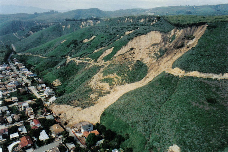 Photograph taken from a helicopter showing a large portion of the vegetation-covered hillside has slid down and buried numerous houses along the base of the slope with a mass of soil and broken bedrock. A light-colored linear feature crosses the hillside descending to the left. This is a pre-existing access road cut into the slope. The road is missing within the landslide deposit, having been disrupted by the downslope movement. Several houses to the left of the landslide are unaffected.