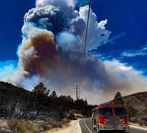 A large smoke column rises thousands of feet above chaparral-covered mountains.